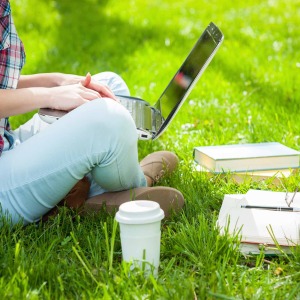 Studying in a green area with laptop, coffee and books stacked on grass area and woman sitting cross-legged